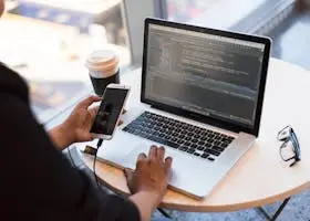 Developer working remotely, coding on a laptop with phone in hand, showcasing modern work culture.